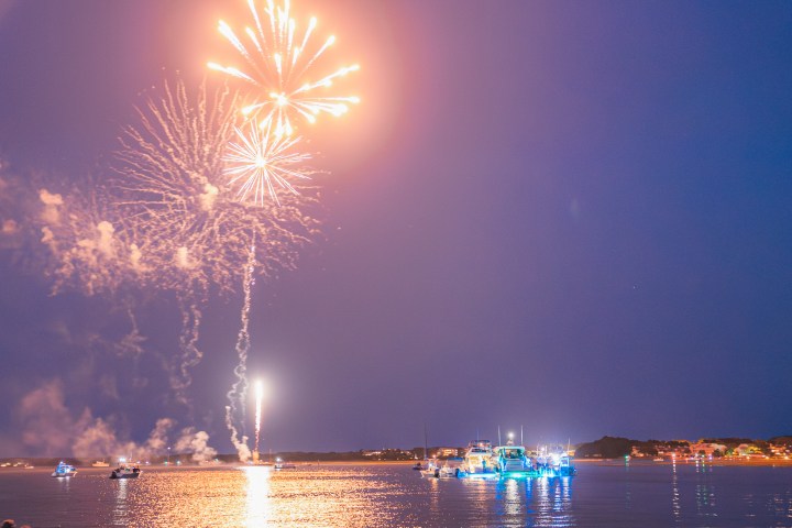 Fireworks exploding over a waterfront with boats and distant city lights at dusk.