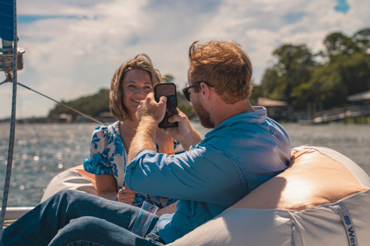 Man taking a photo of a woman on a boat with a sunny sky and water background.